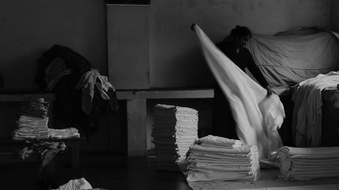 Black and white interior of a dhobi ghat — a worker shakes out a large white sheet beside tall stacks of folded laundry and bundled clothes.