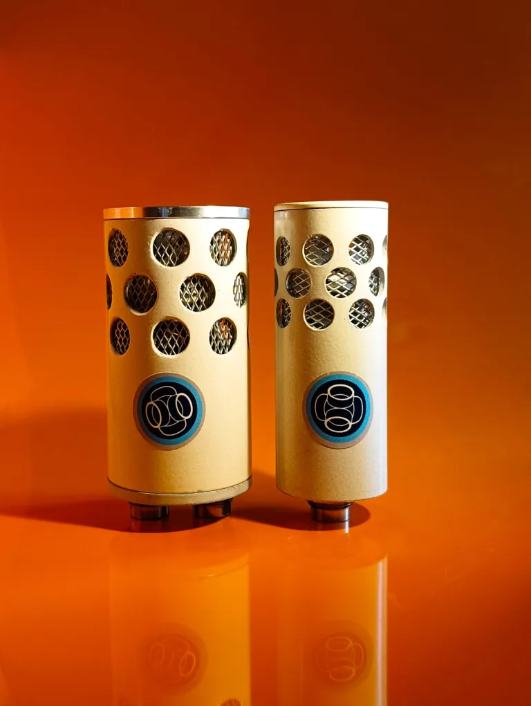 A pair of cylindrical ambisonic microphones with perforated mesh grilles on a warm orange reflective surface.