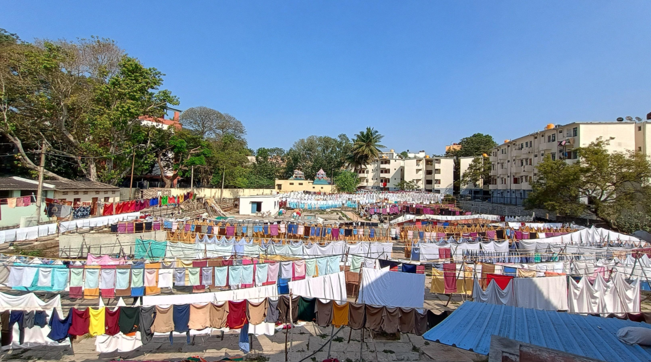 Wide view of a Bangalore dhobi ghat — rows of brightly coloured laundry strung across the open yard under a clear blue sky, framed by trees and apartment blocks.